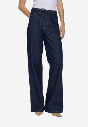 Kobieta nosząca ciemnoniebieskie dżinsy o szerokich nogawkach z wiązanym paskiem w talii, w połączeniu z beżowymi, szpiczastymi butami i białą bluzką.