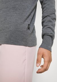 Grauer Strickpullover mit geripptem Saum und Bündchen, mit einem schwarzen Etikett an der Seite. Kombiniert mit hellrosa Hosen.