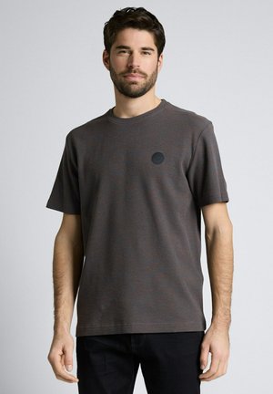 Mann mit kurzen dunklen Haaren und Bart, der ein dunkelgraues, kurzärmeliges, strukturiertes Hemd und schwarze Hosen vor einem einfarbigen Hintergrund trägt.