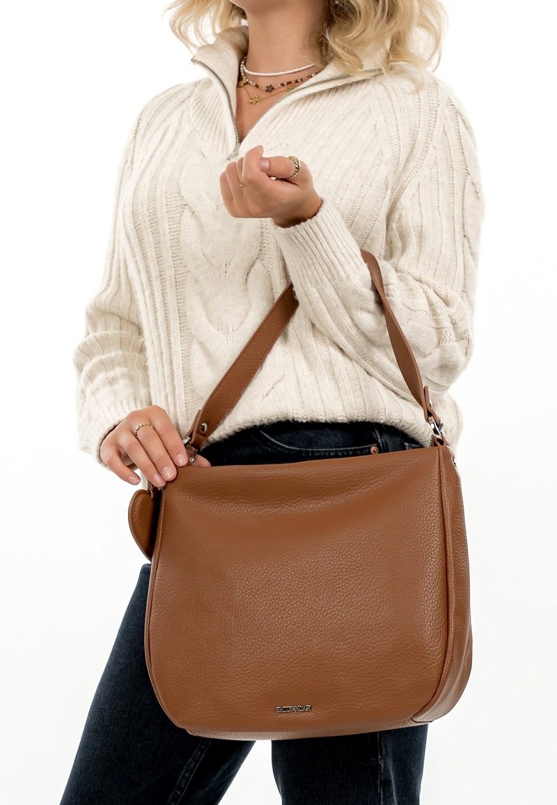Femme portant un pull torsadé crème et un jean noir tenant un grand sac à main en cuir marron avec une bandoulière.