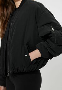 Veste bomber noire avec une finition lisse et brillante, des poignets et une taille côtelés. Comprend des poches zippées et une fermeture à glissière à l'avant.