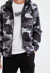 Schwarz-graue Camouflagejacke mit Reißverschluss vorne, elastischen Bündchen und leichtem Material, getragen über einem weißen T-Shirt mit dem Schriftzug "NOP".
