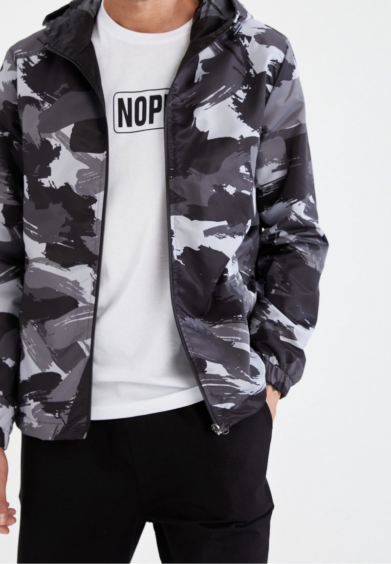 Schwarz-graue Camouflagejacke mit Reißverschluss vorne, elastischen Bündchen und leichtem Material, getragen über einem weißen T-Shirt mit dem Schriftzug "NOP".