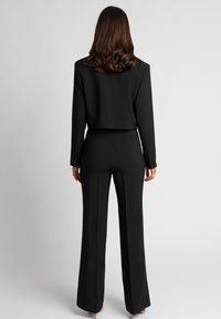 Czarny garnitur na miarę z krótką marynarką i spodniami o szerokich nogawkach. Gładka tkanina o czystych liniach i dopasowanym kroju. Mankiety zapinane na guziki.