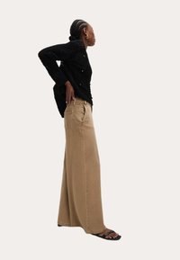 Pull en tricot noir texturé avec boutons associé à un pantalon large beige. Modèle portant des sandales plates noires, en position de profil.