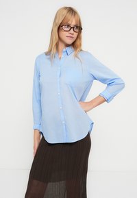 Chemise bleu clair en coton, avec une poche poitrine, des manches longues retroussées aux poignets et un ourlet arrondi.
