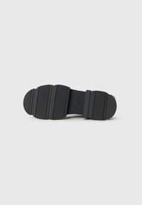 Semelle de chaussure noire avec une texture antidérapante et trois larges rainures horizontales sur les sections du talon et des orteils, sur un fond blanc uni.