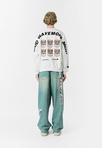 Sudadera blanca con estampado gráfico que presenta latas en la parte posterior. Jeans de pierna ancha, de color azul claro, con textura desgastada y etiquetas visibles. Zapatos con suela de goma.