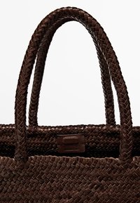 Dunkelbraune geflochtene Lederhandtasche mit zwei runden Griffen und einem kleinen rechteckigen Markenlabel, das nahe der oberen Kante aufgenäht ist.