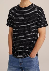 Man met een zwart T-shirt met korte mouwen met witte horizontale streeppatronen, gecombineerd met blauwe denimjeans, handen in de zakken.