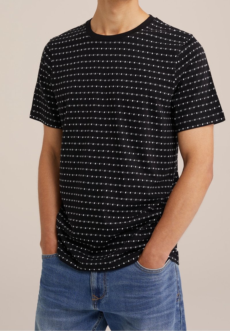 Man met een zwart T-shirt met korte mouwen met witte horizontale streeppatronen, gecombineerd met blauwe denimjeans, handen in de zakken.