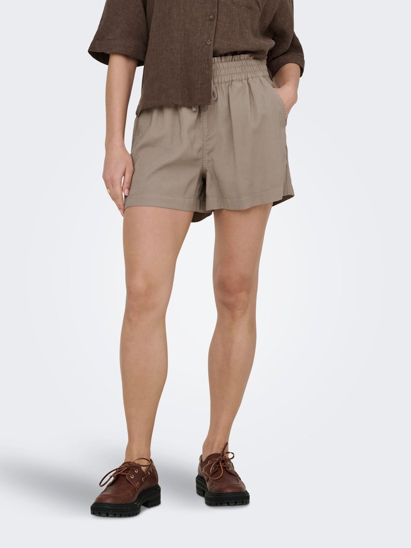 Kvinde iført beige elastiske shorts med talje og brune snørebåndssko, der står med den ene hånd i lommen mod en ensfarvet baggrund.