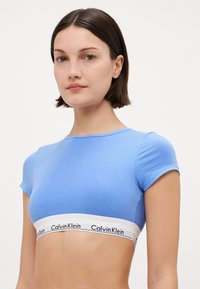 Šviesiai mėlynas trumpas viršus su trumpomis rankovėmis ir baltu elastiniu juostele, kurioje yra "Calvin Klein" prekės ženklas. Minkštas, prigludęs dizainas, minimalus tekstūros pojūtis.