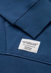 Felpa blu con colletto e polsini a costine, caratterizzata da un'etichetta bianca che riporta la scritta "IL TUO UNICO LIMITE È LA TUA MENTE" e "OMBRE dal 1985."