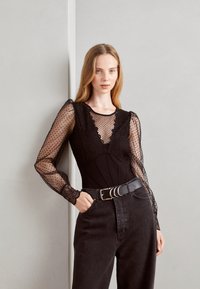 Body nero con maniche lunghe trasparenti a pois e dettagli in pizzo. Abbinato a jeans scuri a vita alta e una cintura nera larga.