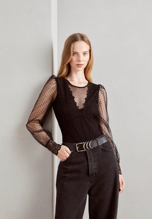 Schwarzer Overall mit transparenten, gepunkteten langen Ärmeln und Spitzenverzierungen. Kombiniert mit hochgeschnittenen dunklen Jeans und einem breiten schwarzen Gürtel.
