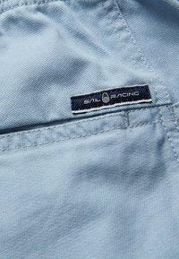 Světle modrá látka s hladkou texturou má přišitou tmavě modrou obdélníkovou etiketu, na které je zobrazeno logo "SAIL RACING".