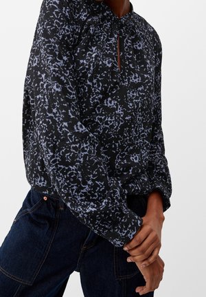 Person iført sort og blå abstrakt mønstret bluse med lange ærmer, stukket ned i mørkeblå denim bukser, hænder blidt foldet.