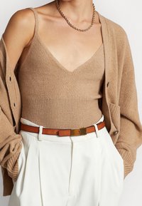 Tan stickad linne med v-ringning, i kombination med en beige kavaj och vita byxor. Outfiten inkluderar ett brunt bälte med en guldfärgad spänne.