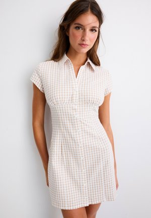Femme aux longs cheveux bruns portant une robe à manches courtes à carreaux beige et blanche boutonnée, sur un fond blanc uni.
