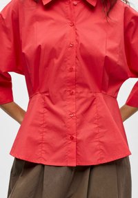 Femme portant une chemise rouge cintrée à boutons avec manches trois-quarts et une jupe marron, les mains sur les hanches, sur un fond blanc.