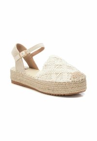 Sandal de esparto tejido en color beige con parte superior texturizada, correa ajustable en el tobillo y tacón de cuña envuelto en yute. Plantilla suave en tono marrón.