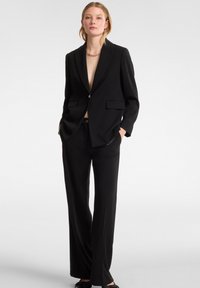 Traje negro a medida que presenta un blazer de un solo botón con solapas anchas, bolsillos frontales dobles y pantalones holgados. Textura de tejido suave.