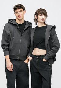 Hanorace gri cu fermoar și buzunare frontale, cu un imprimeu subtil cu logo. Purtate peste bluze negre, asortate cu blugi închiși la culoare, largi.