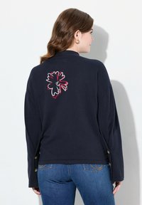 Mörkblå långärmad topp med blommig broderi i vitt och rött på baksidan, med knappr förslutningar på ärmarna. Strukturerad tyg.