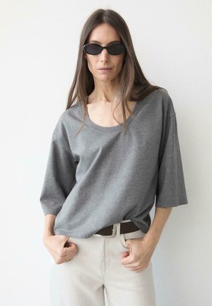 Kobieta o długich brązowych włosach, nosząca czarne owalne okulary przeciwsłoneczne, luźny szary T-shirt oraz jasne spodnie z ciemnym paskiem, pozująca na tle białej ściany.