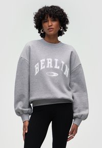 Szary sweatshirt z białym napisem "BERLIN", z opadającymi ramionami i objętościowymi rękawami, w połączeniu z czarnymi prążkowanymi legginsami.