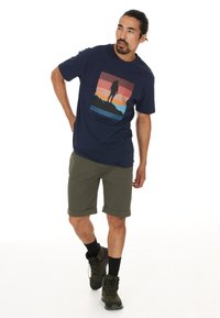 Mörkblå bomullströja med grafisk design som visar lager av solnedgångsfärger och en silhuett av en vandrare. Kombinerad med uppvikta olivgröna shorts.