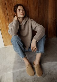 Sabots en daim tan avec un détail de boucle, portés avec un jean bleu clair retroussé et un hoodie beige. Texture lisse sur un fond minimaliste.