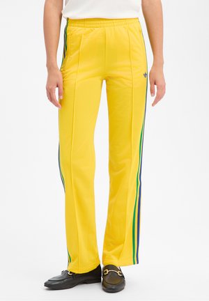 Pantalon de sport jaune avec des rayures latérales bleues et vertes, coupe ajustée, porté avec des mocassins noirs et un haut blanc.