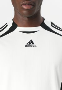 Balta sporta krekls ar vertikālu ribotu tekstūru, melniem akcentiem uz apkakles un pleciem, ar melnu izšūtu Adidas logo.