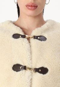Outerwear in shearling color crema con chiusure in pelle marrone scuro, dettagli in hardware dorato e una finitura morbida e testurizzata.