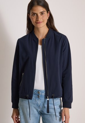 Femme portant une veste zippée bleu marine sur une chemise blanche et un jean bleu clair, debout devant un fond clair uni.