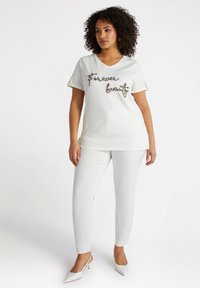 Camisa blanca de manga corta y escote en V con un adhesivo texturizado que dice "Para siempre hermosa", combinada con pantalones ajustados blancos y tacones de punta.