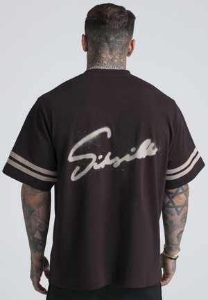 Uomo con tatuaggi sul collo e sulle braccia che indossa una maglietta nera a maniche corte con strisce beige sulle maniche e scritte beige in stile spray sul retro.