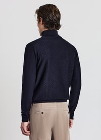 Marineblauer Strickpullover mit hohem Kragen, strukturiertem Rautenmuster, gerippten Bündchen und Saum. Kombiniert mit hellen khakifarbenen Hosen.