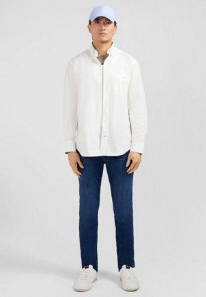 Chemise blanche à manches longues avec des détails de boutons, jeans bleu foncé et sneakers blanches. Les accessoires incluent une casquette bleu clair. Design simple et décontracté.