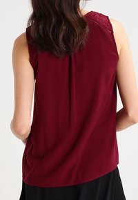 Burgundy sleeveless top with a textured pattern. Features a lace detail at the neckline and a curved hem at the back. Soft fabric.