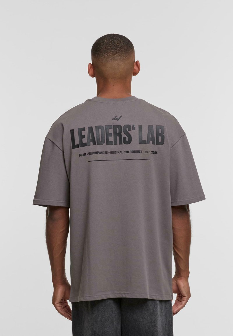 Szara, przejrzysta koszulka wykonana z bawełny. Na plecach widnieje napis "LEADERS' LAB" w wielkim czarnym druku, z dodatkowymi oznaczeniami poniżej.