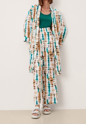 Personne portant un pantalon ample à motifs beige, turquoise et marron, avec une veste longue assortie ouverte, un débardeur vert foncé et des sandales à semelles compensées blanches.