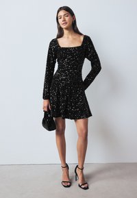 Robe à sequins noire avec des manches longues, décolleté carré, taille ajustée et jupe évasée. Assortie à des talons noirs à lanières et à un petit sac à main.