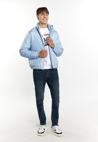 Světle modrá péřová bunda se zipovým límcem, nošená přes bílou mikinu, v kombinaci s tmavými džíny a bílými teniskami.