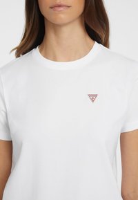 Hvit T-skjorte i bomull med korte ermer, med en rød, trekantet logo på venstre bryst. Glatt tekstur og klassisk design.