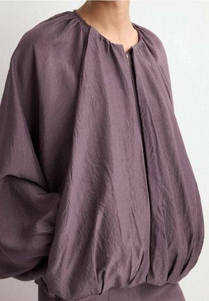 Blouse violette ample avec encolure froncée et manches ballon volumineuses sur une personne devant un fond clair uni.