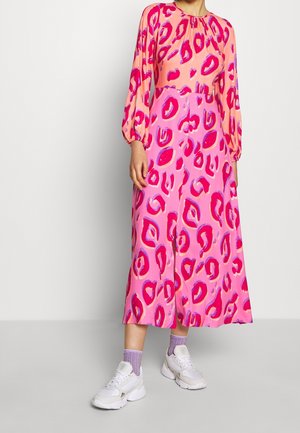 Kvinde iført en lang pink og ferskenfarvet kjole med røde og lilla abstrakte pletter, kombineret med hvide sneakers og lavendelfarvede netstrømper.