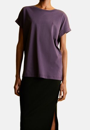 Fioletowy T-shirt z krótkim rękawem i podwiniętymi mankietami, o luźnym kroju, z małym logo. Dobraną do niego czarną spódnicę z bocznym rozcięciem.